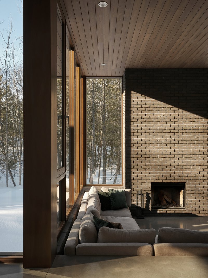 Living room with timber ceiling and brick fireplace wall framed by tall windows overlooking winter forest