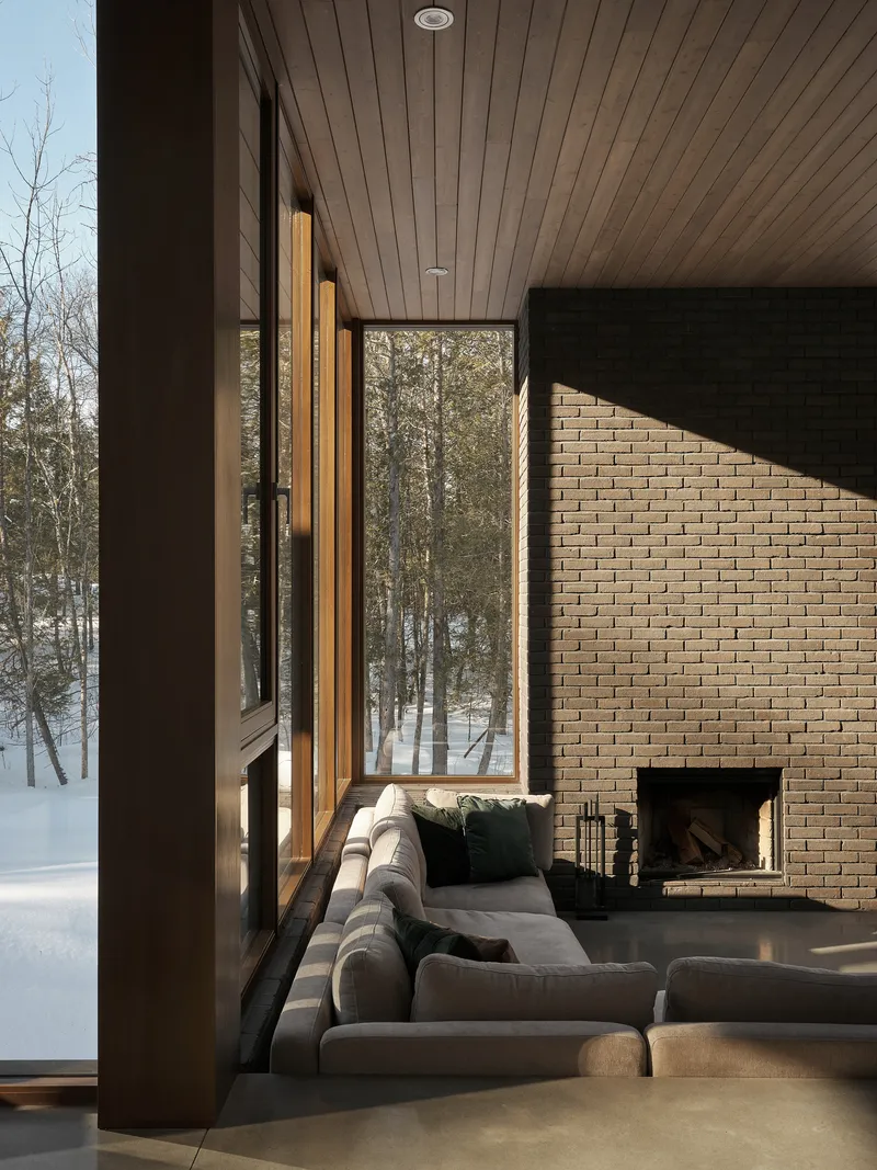 Living room with timber ceiling and brick fireplace wall framed by tall windows overlooking winter forest