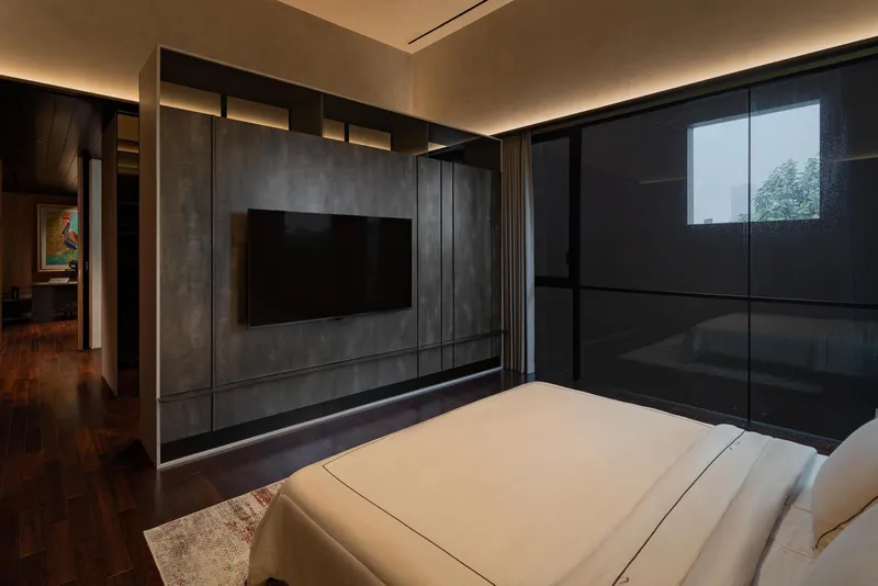 Bedroom corner showing dark paneled wall with recessed television and floor-to-ceiling glazed corner opening to greenery