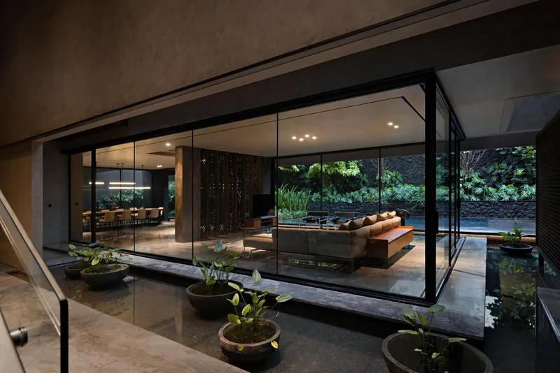 Glass-enclosed living pavilion suspended over reflecting pool courtyard with planted beds and steel handrail stair at evening