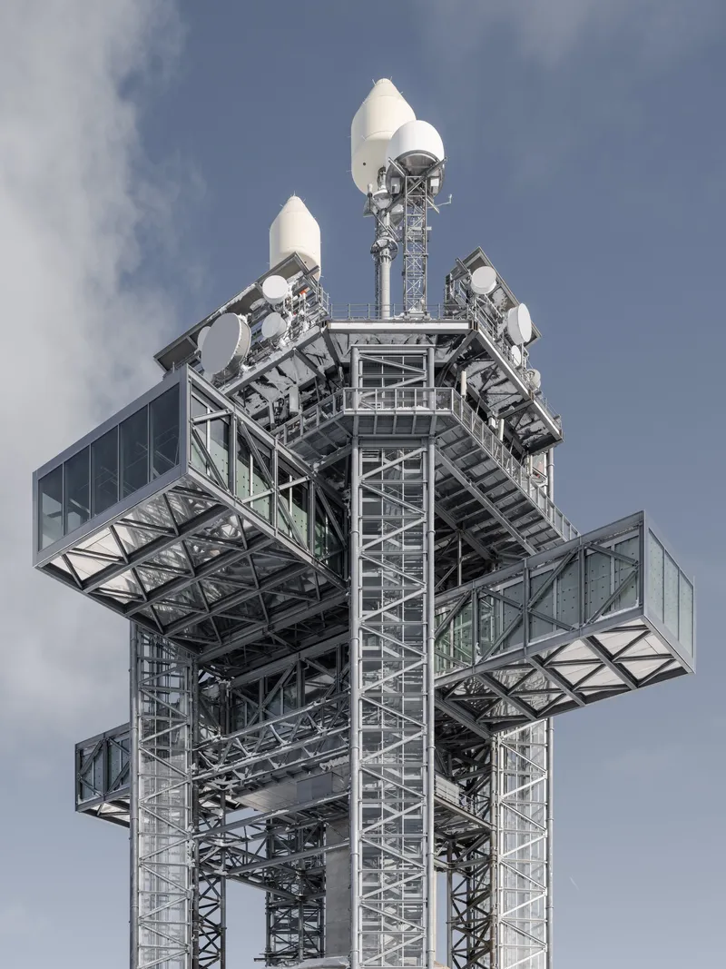 Close-up of the tower top showing exposed steel trusses, glazed platforms, and white radome equipment