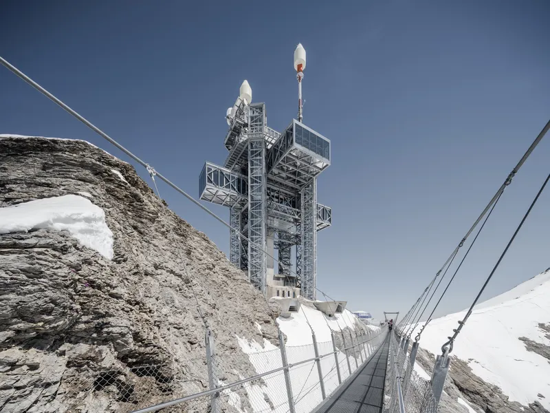 Steel-framed tower structure rising above a suspension bridge spanning exposed rock and snow