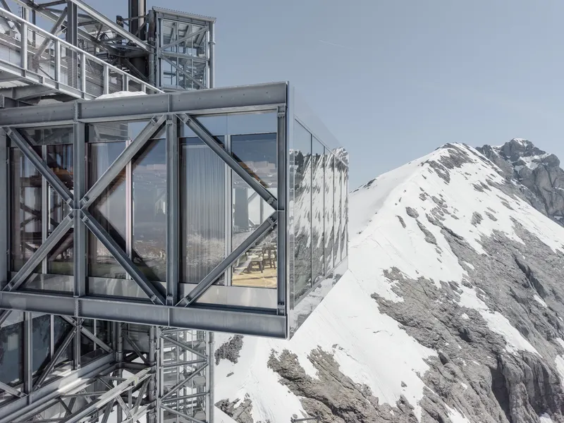 Glass-enclosed viewing platform cantilevered from the steel frame with mountain peaks beyond