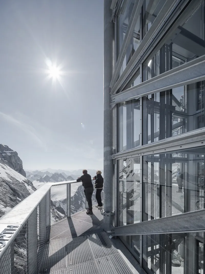 Two visitors on the exterior metal walkway beside the glazed tower under bright sun