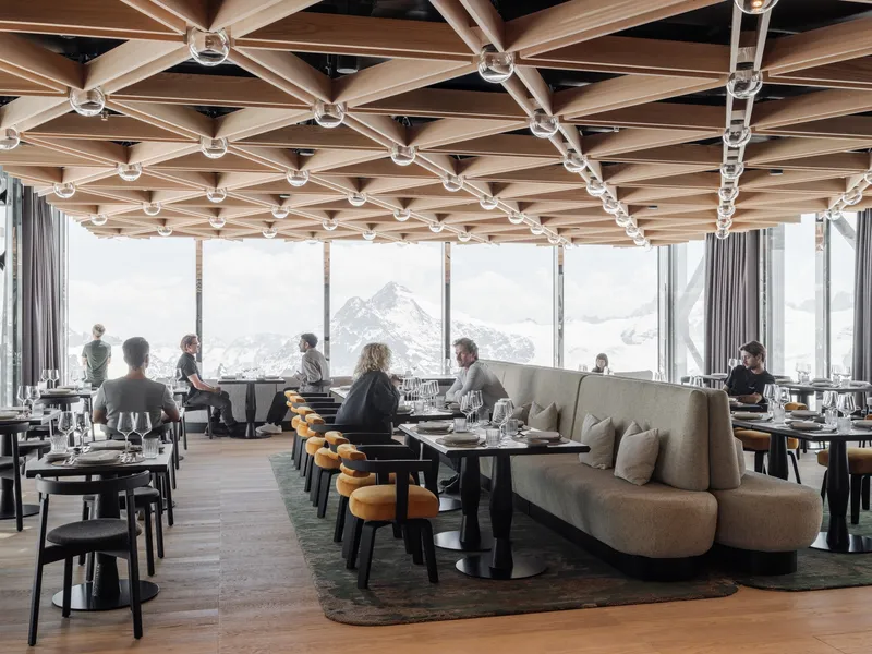 Restaurant interior with geometric timber ceiling grid and floor-to-ceiling windows overlooking snow peaks