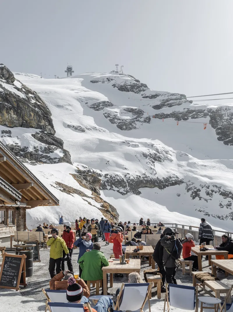 Outdoor terrace with timber seating beneath a timber-framed canopy facing snow-covered mountain peaks and gondola cables