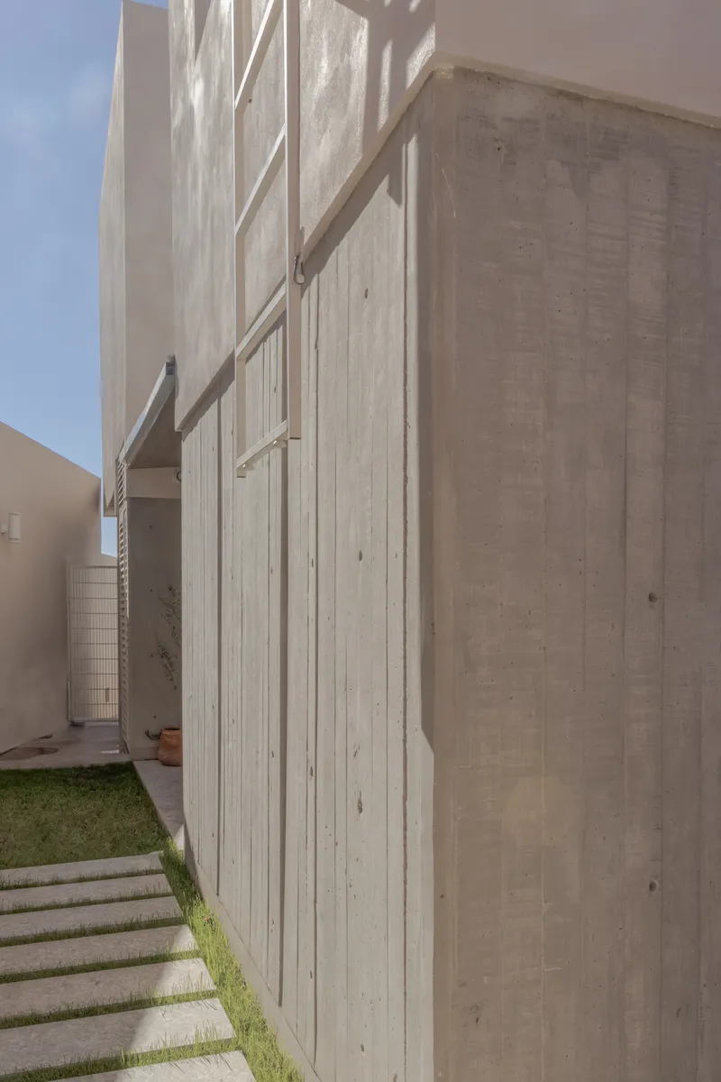 Boardmarked concrete facade with projecting horizontal louvers beside a grass pathway in sunlight