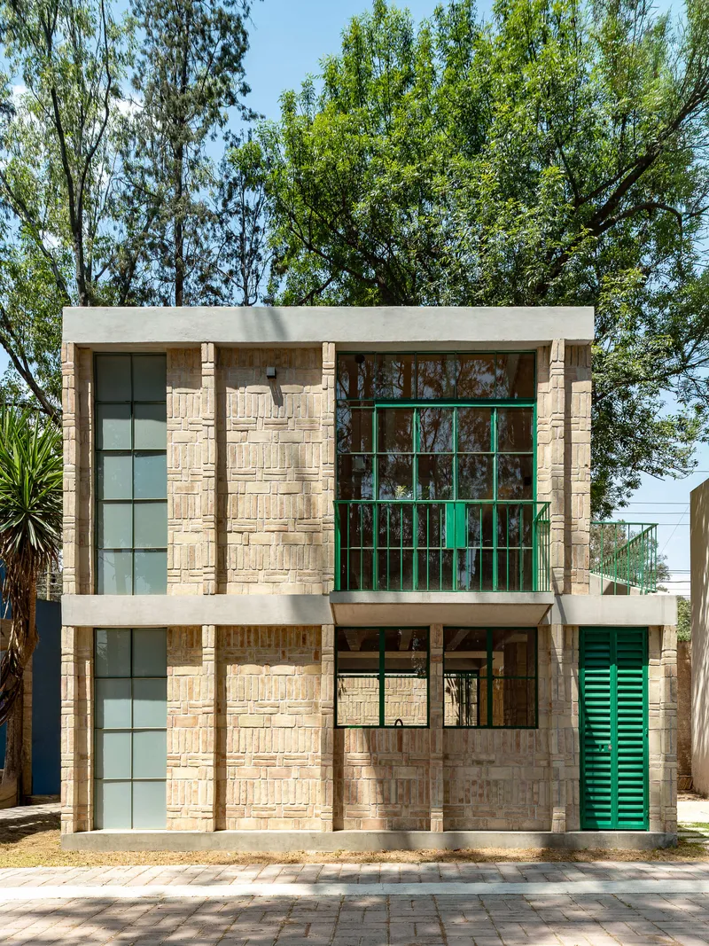 Street facade with textured brick walls, green metal-framed windows, and concrete bands framed by tall trees