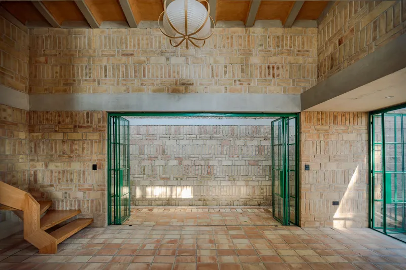 Interior view through open green-framed doors to textured brick courtyard wall with exposed timber ceiling beams