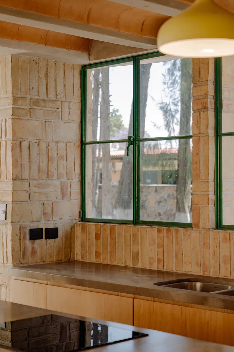 Kitchen counter with green-framed window in textured brick wall under timber ceiling and pendant light