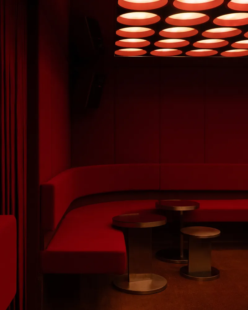 Red pleated curtain beside curved banquette seating with bronze stools under the glowing ceiling grid