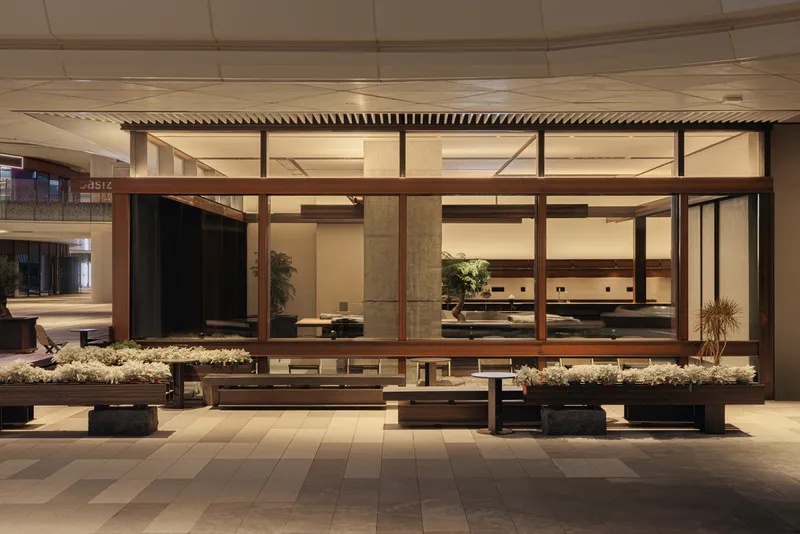 Glass-fronted coffee pavilion within a tiled indoor plaza with planted beds flanking timber benches