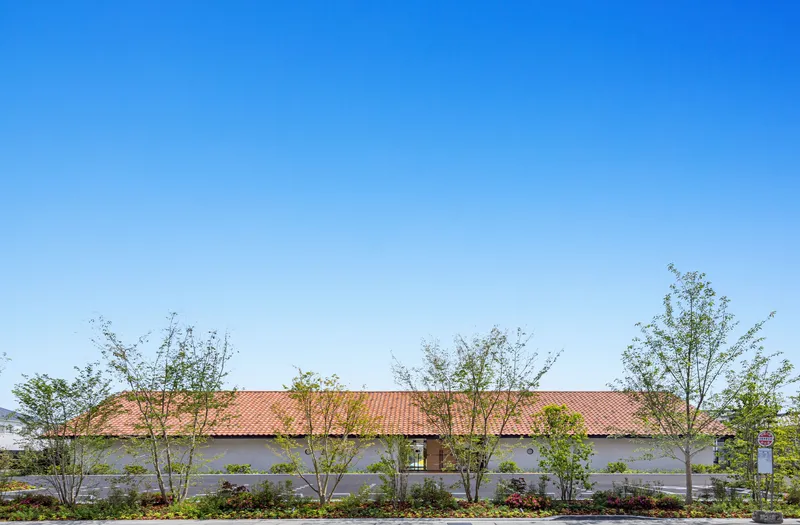 Long elevation showing the red tile roof with white stucco walls and young trees planted along the perimeter