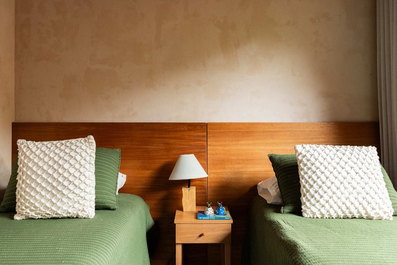 Twin beds with timber headboard and central nightstand beneath wall-mounted reading lamp in soft daylight