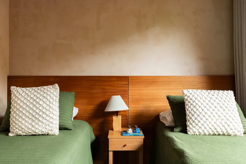 Twin beds with timber headboard and central nightstand beneath wall-mounted reading lamp in soft daylight