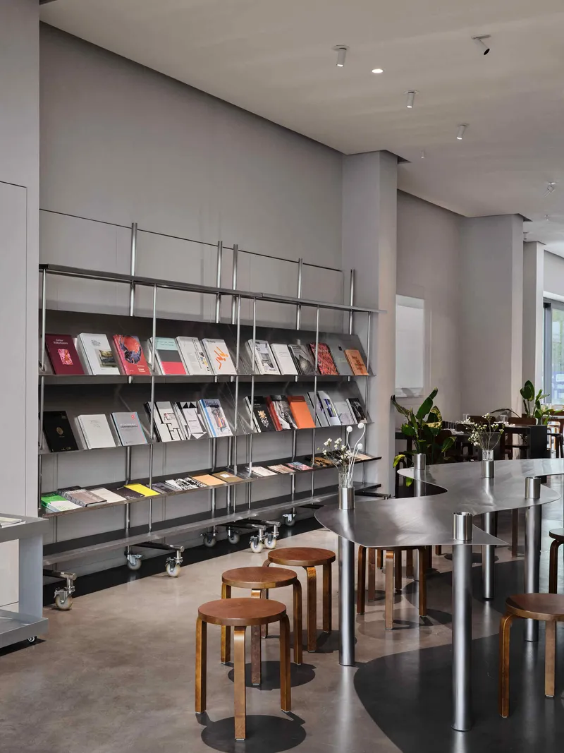 Steel magazine rack with angled shelves beside timber stools and metal tables under track lighting