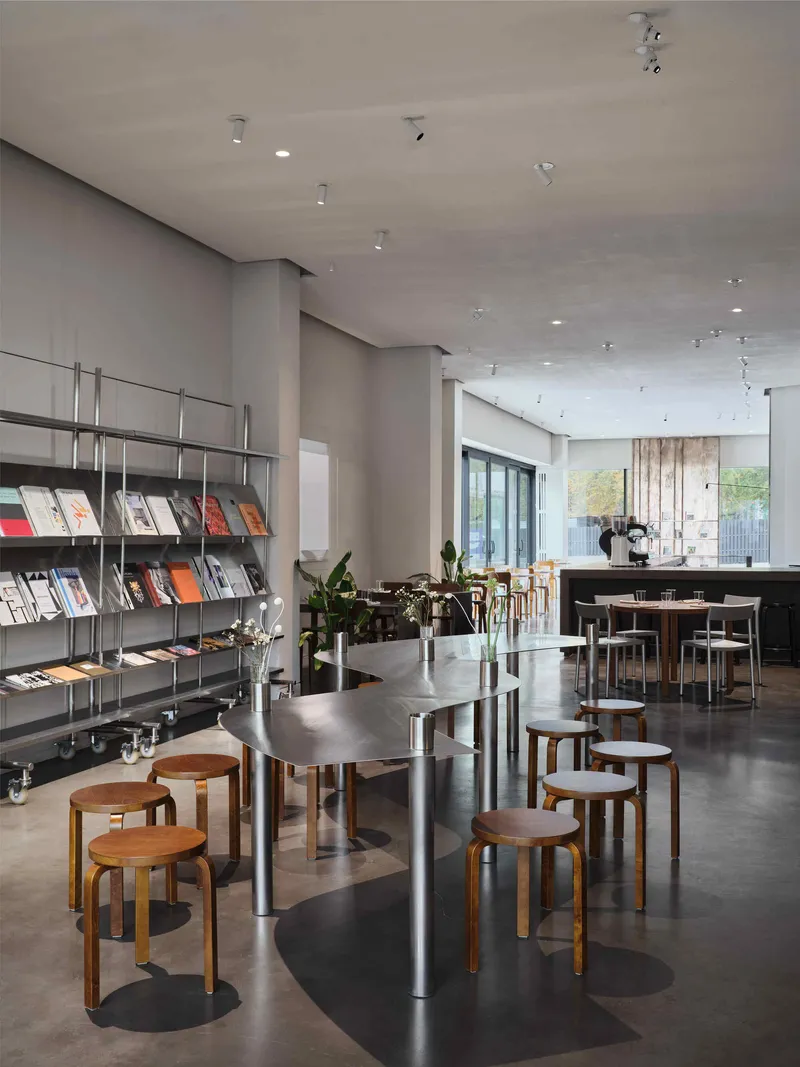 Open cafe space with magazine shelving, timber stools, and daylight through glazed doors beyond potted plants