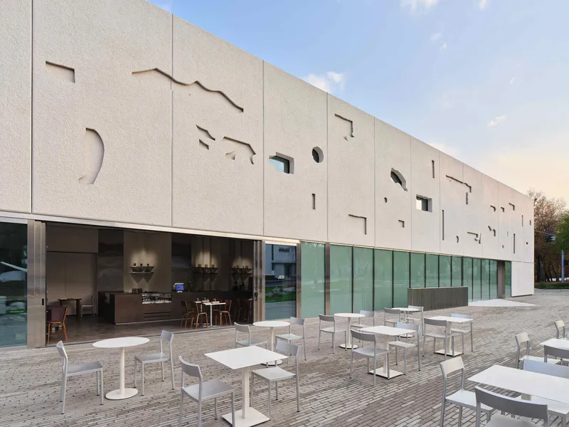 Exterior facade with cut-out patterns above glass storefronts and white outdoor seating on a cobbled terrace