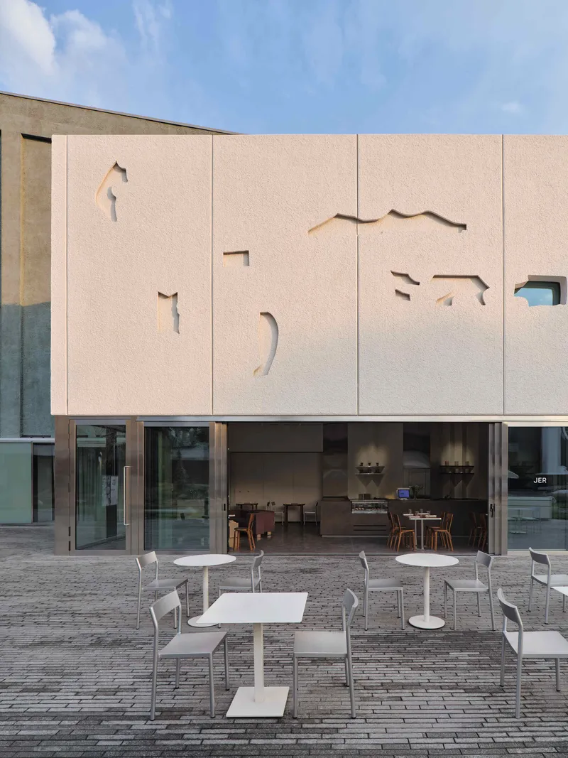 Pale facade with cutout animal shapes above a glass-walled cafe terrace with white outdoor furniture