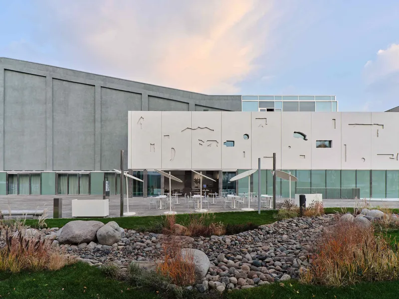 Cantilevered facade with organic cutouts above a glazed base, adjacent to a landscaped rock garden at dusk