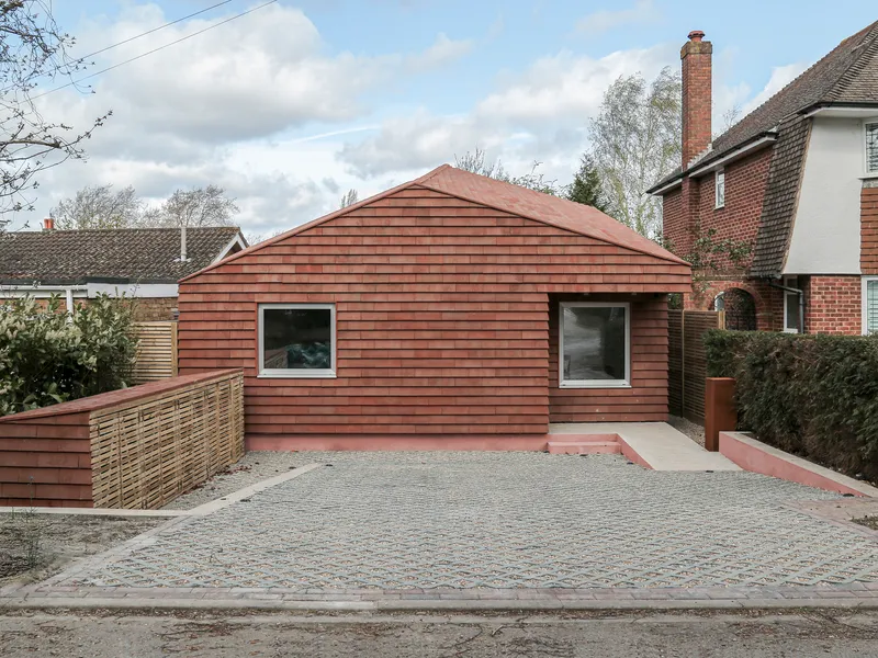 Front view of the terracotta tile-clad volume with two square windows and permeable paving under cloudy skies