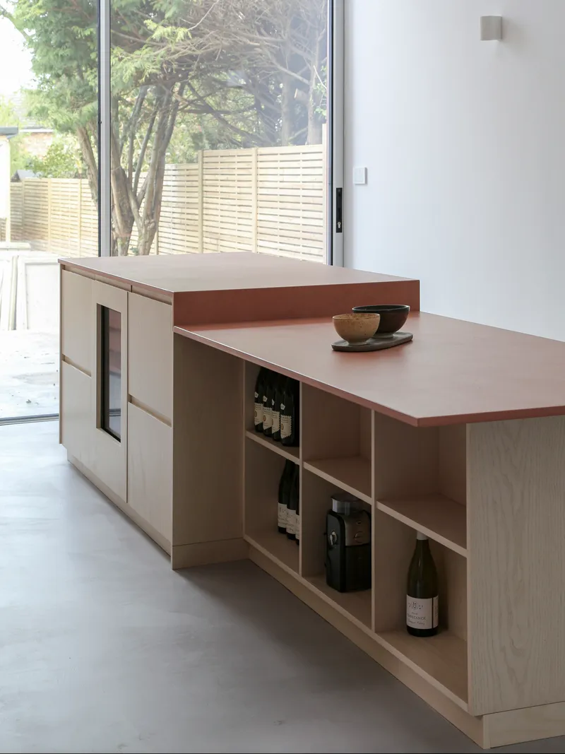 Kitchen island with open shelving for wine storage and view to horizontal slatted fence beyond glazing