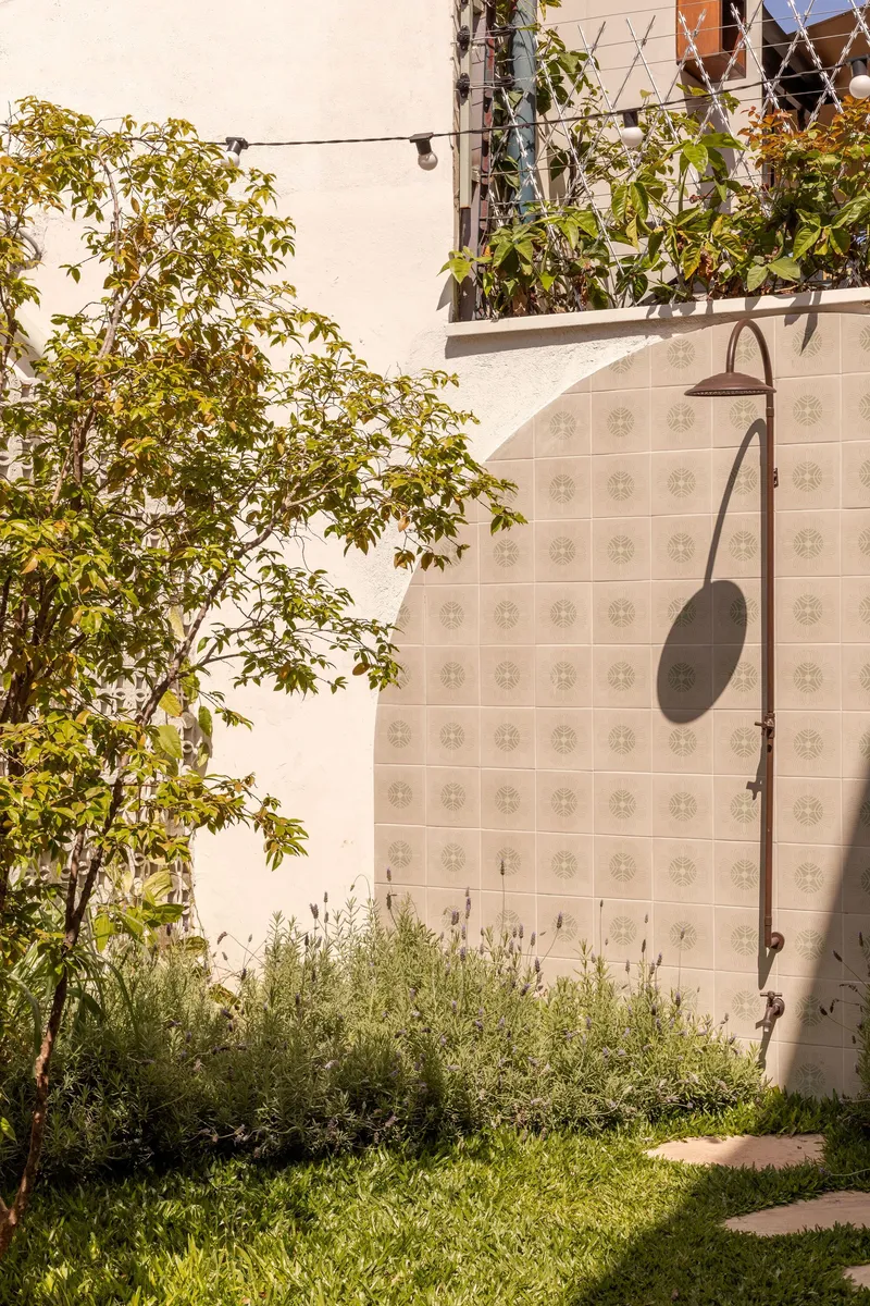 Outdoor shower fixture on patterned tile wall beside planted tree and grass in afternoon sun