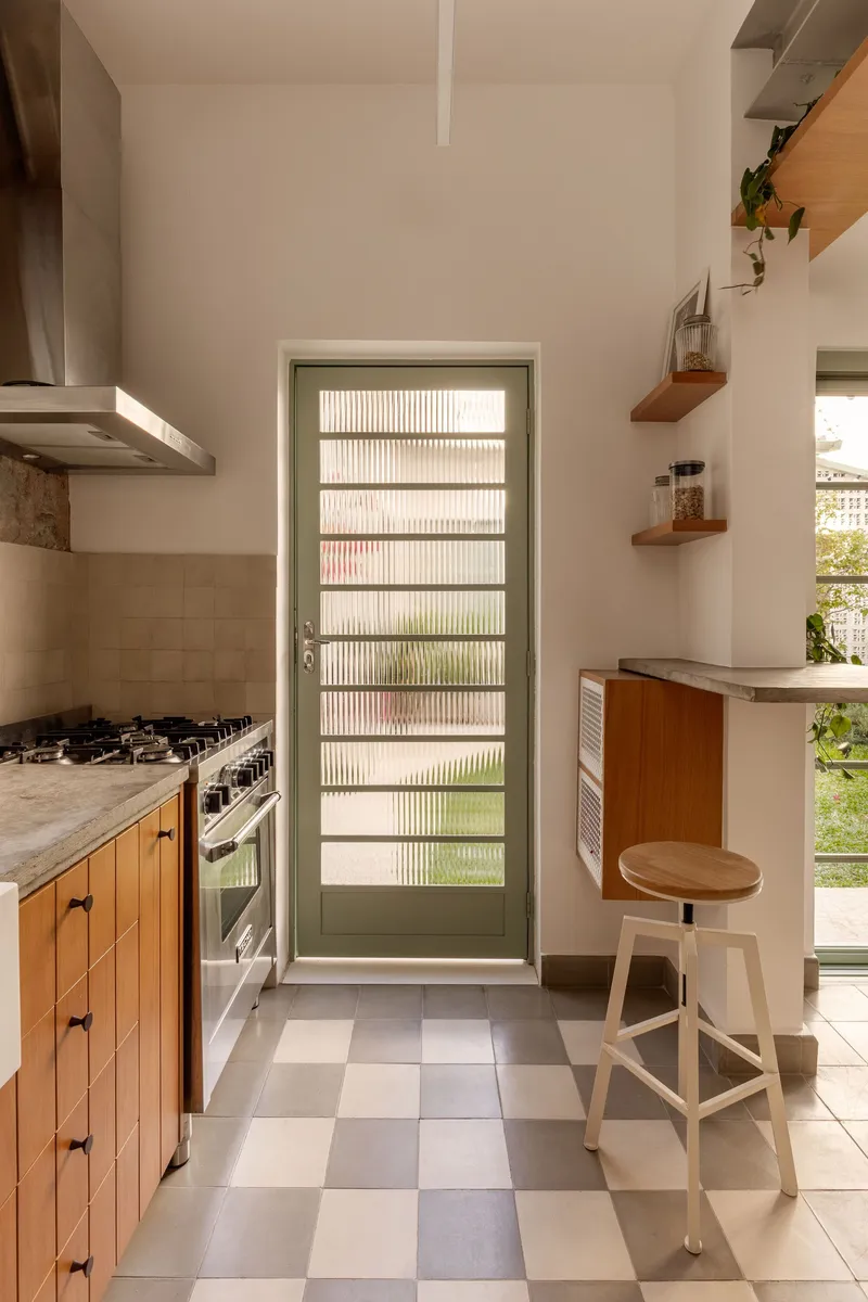 Narrow kitchen passage with checkered floor, timber cabinetry, green-framed glass door, and floating timber shelves