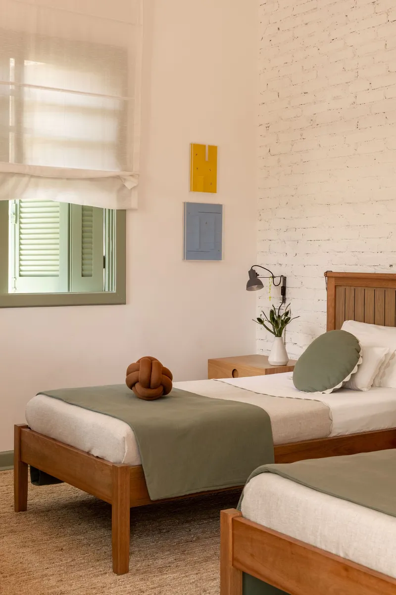 Twin beds with sage green runners beneath whitewashed brick wall and window with Roman shade