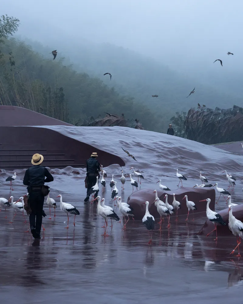 Two visitors observing white ibises on wet corten plaza under misty mountain backdrop