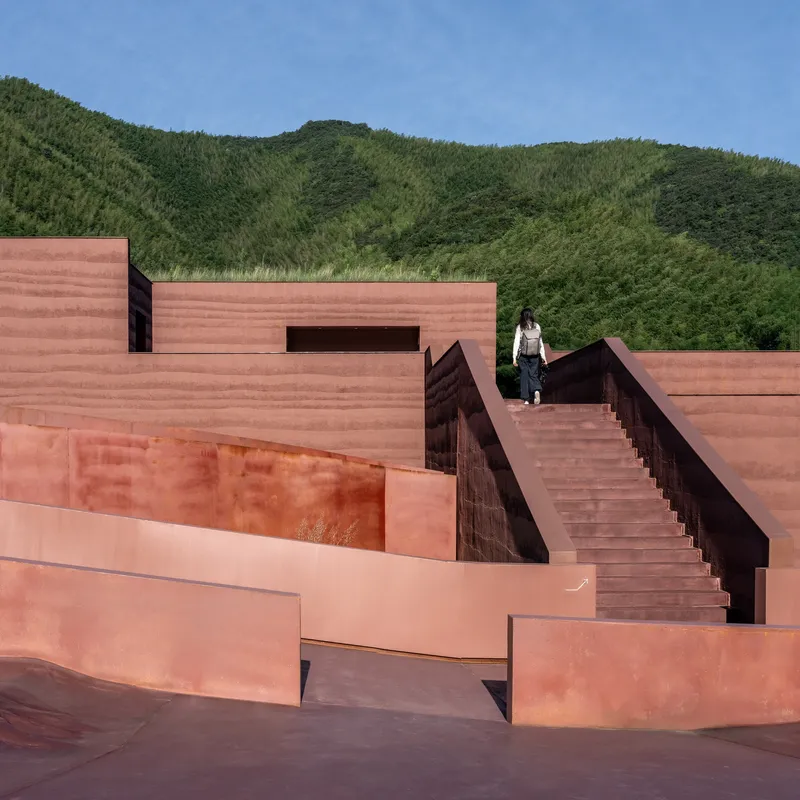 Person ascending a diagonal staircase between red rammed earth walls and hillside beyond
