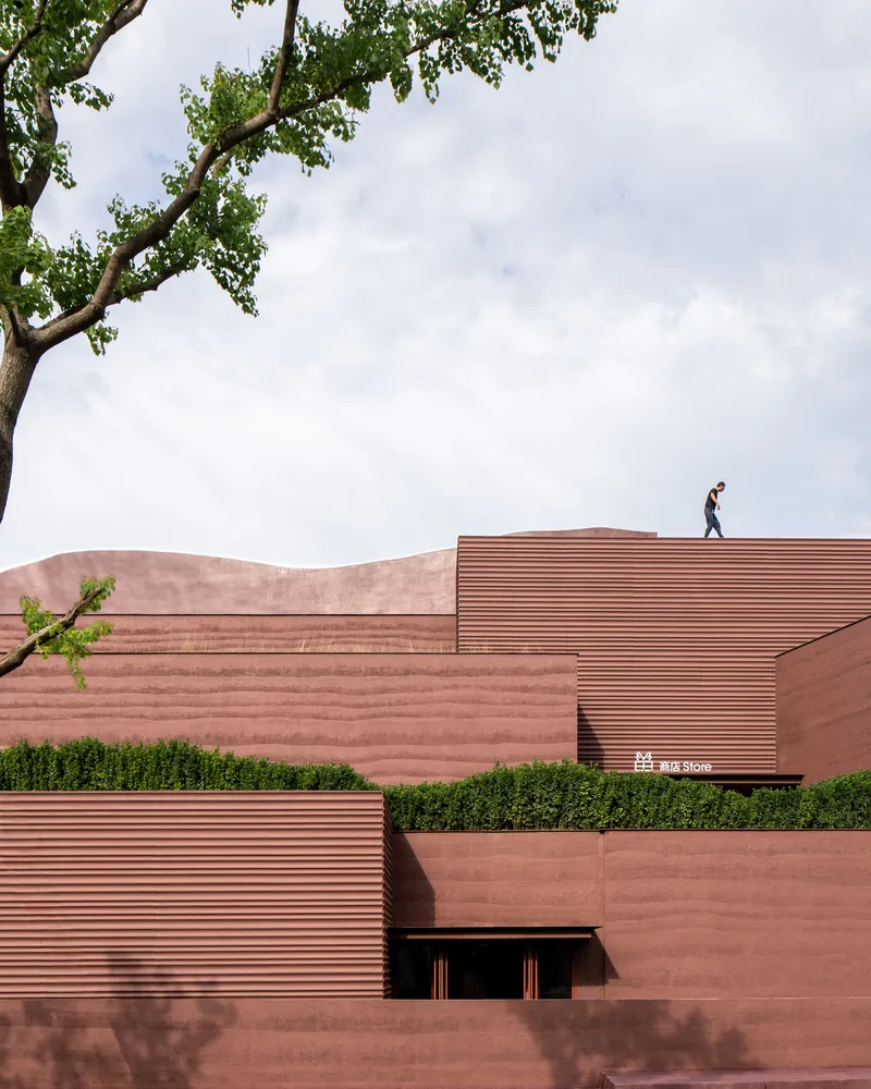 Layered red stone facades with horizontal ribbing and a person walking the roofline under tree branches