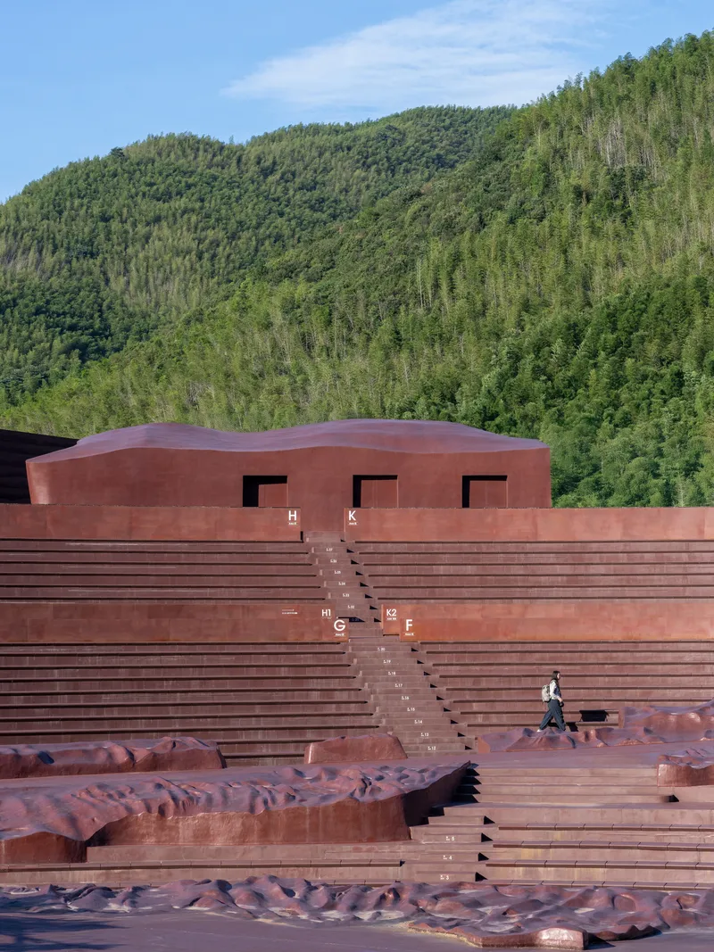Terraced amphitheater with weathered steel seating and a curved roof volume against a forested hillside