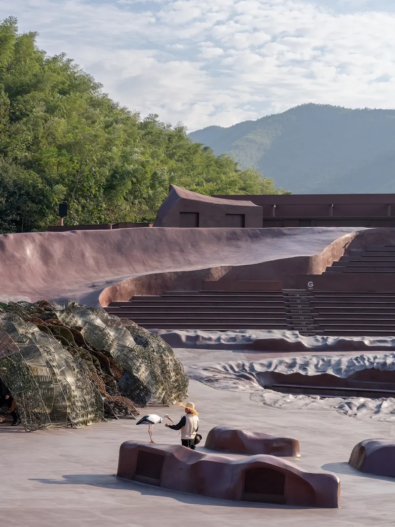 Outdoor courtyard with sculptural seating elements and a visitor beneath cloudy skies and distant peaks