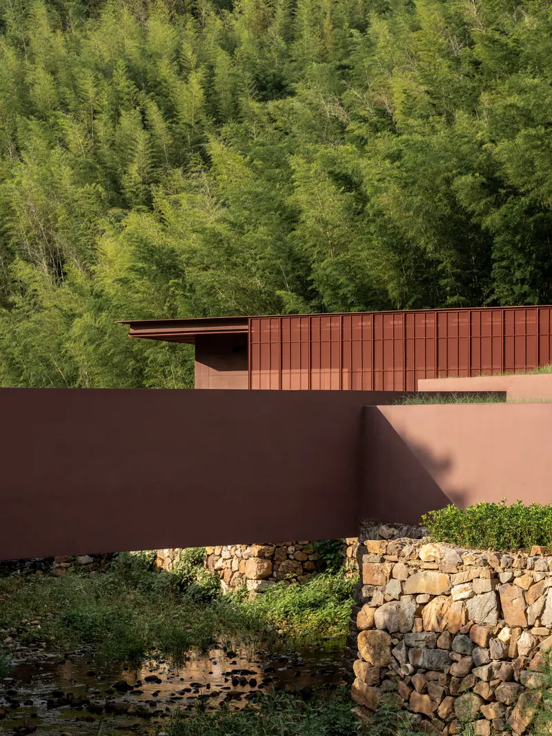 Corten steel bridge crossing a stone retaining wall and stream against a dense bamboo forest