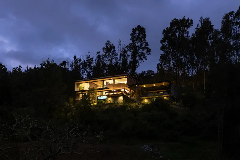 Illuminated residence nestled into forested hillside under blue hour sky with eucalyptus silhouettes