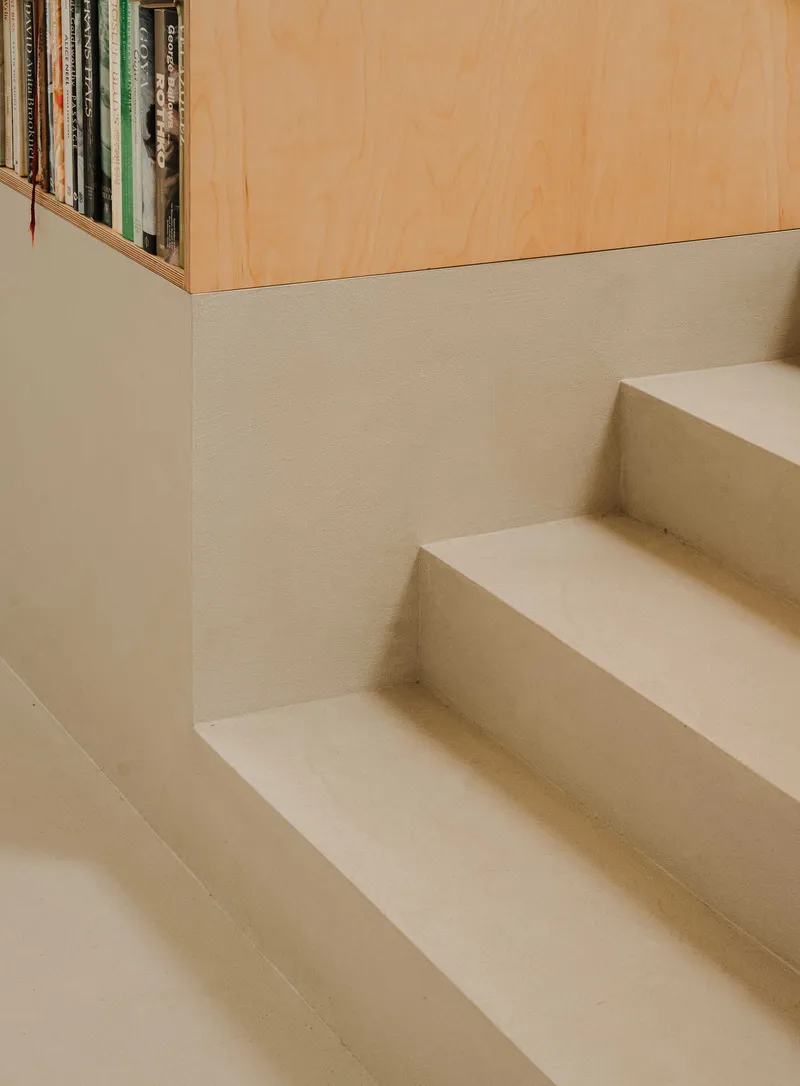Detail of plaster staircase treads meeting a white wall beneath a plywood bookshelf