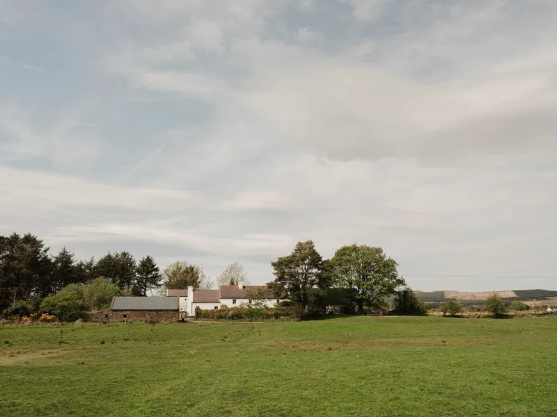 Low whitewashed farmhouse in a grass field with mature trees under an overcast sky