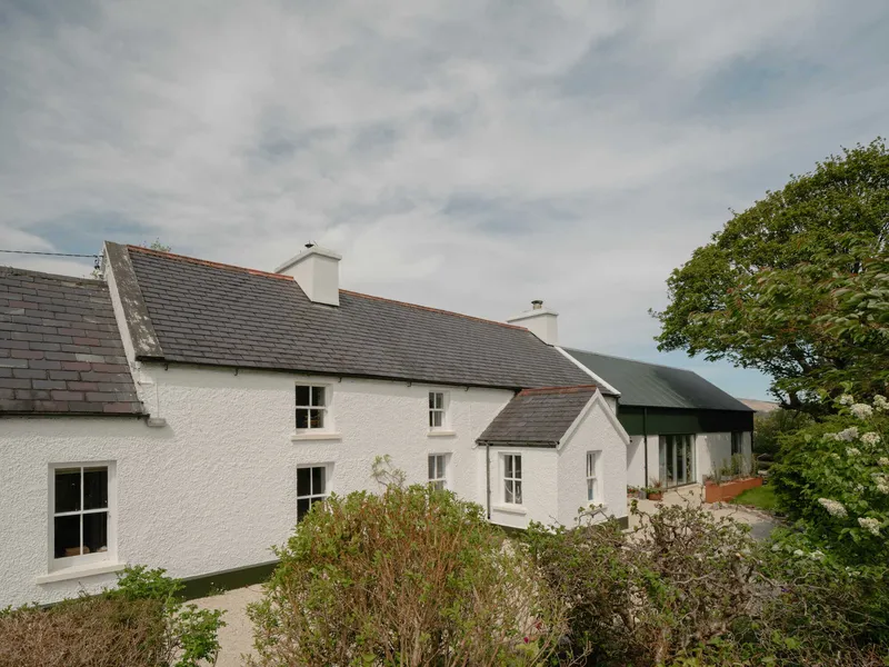 Whitewashed cottage with grey tiled roofs and a green corrugated metal extension under cloudy skies