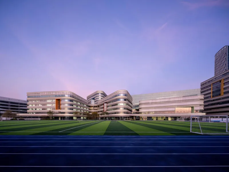 View across the sports pitch toward the banded facades and curved volumes at dusk