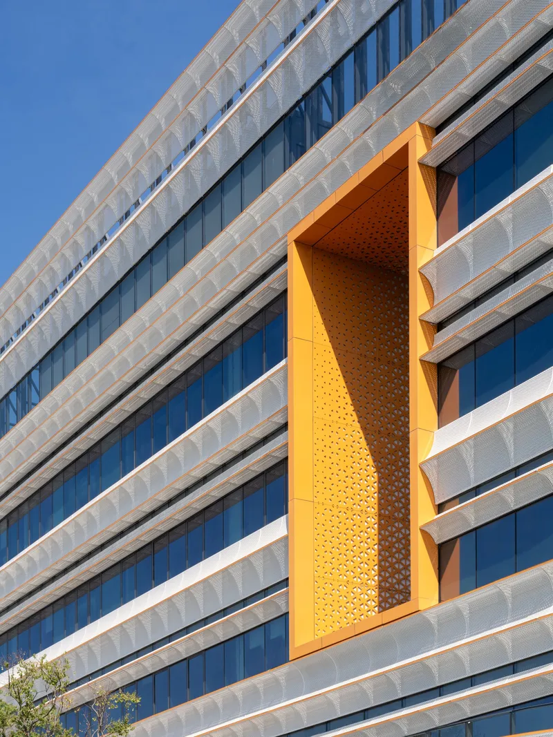 Detail of perforated orange metal panel set into the horizontal banded facade under blue sky