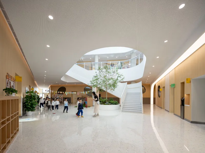 Interior atrium with spiraling white staircase, circular skylight above, and children gathering beneath a planted tree