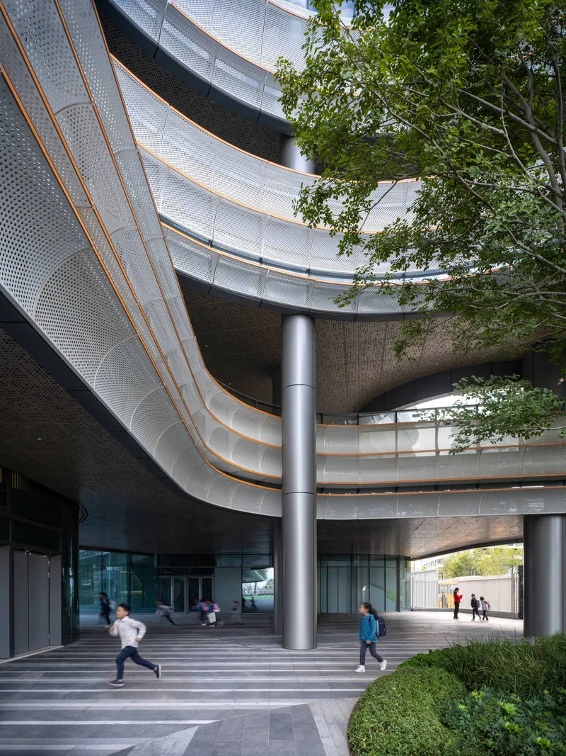 Covered entry plaza with sweeping perforated metal canopy, cylindrical columns, and children running past planted beds