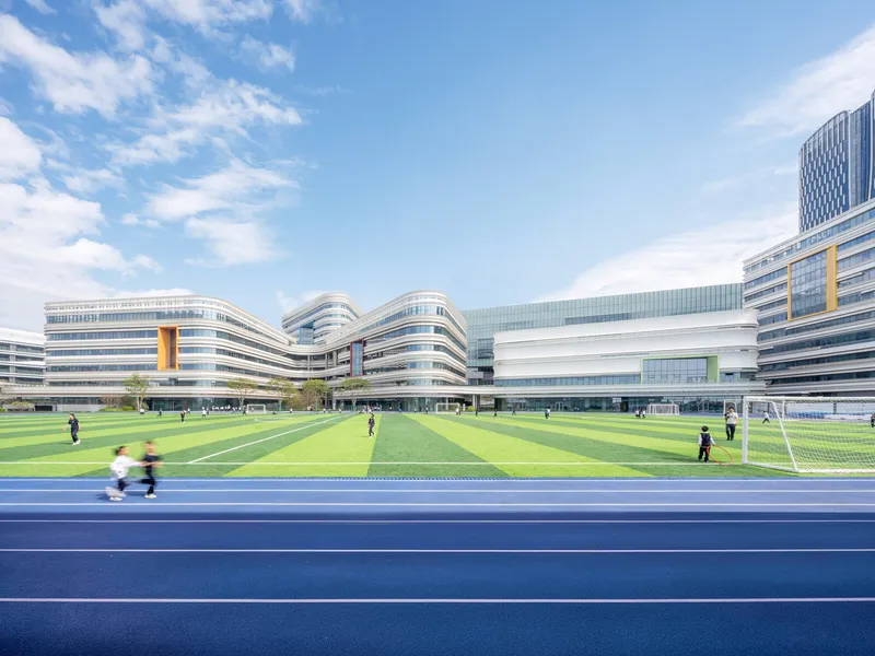 View across the blue running track and green football pitch toward the curved horizontal facades under scattered clouds