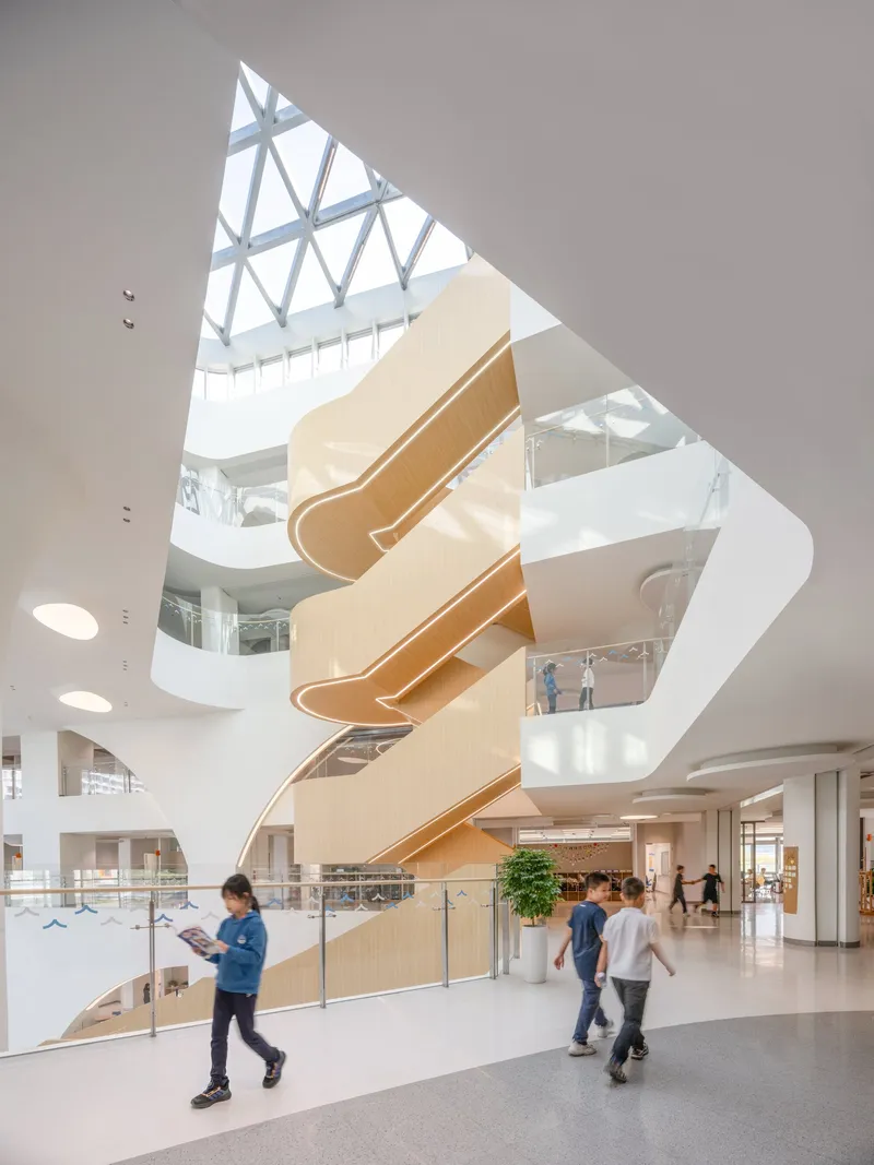 Central atrium with spiraling timber staircase beneath a triangular skylight as students pass through the lobby