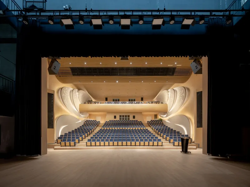 View from the stage into the auditorium showing curved balconies, timber acoustic ceiling, and rows of blue seats
