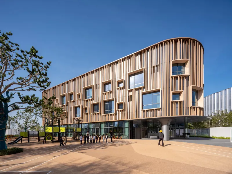 Vertical timber screen facade with irregular window openings and people walking across the plaza in afternoon light