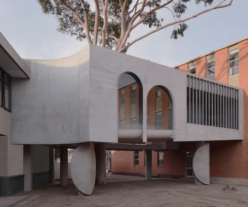 Elevated concrete volume on circular legs with arched openings framing an existing brick building