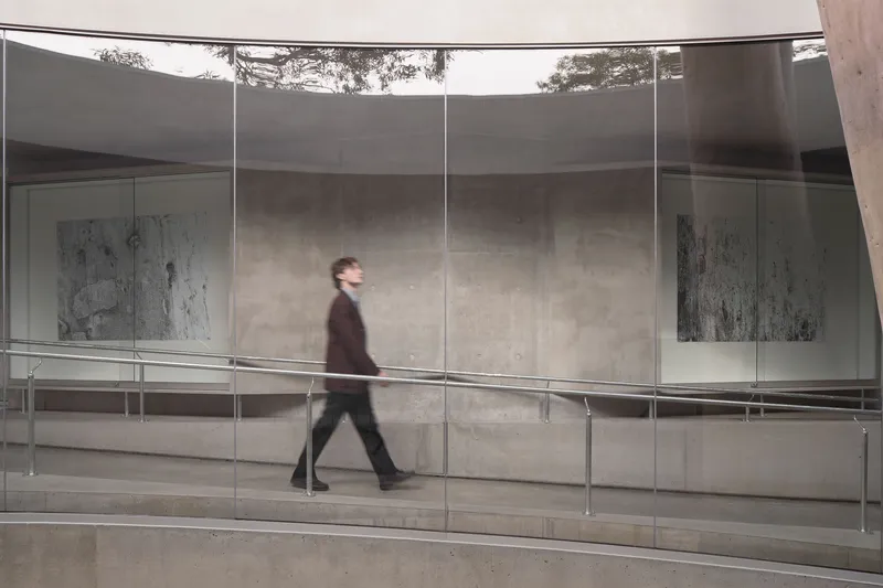 Glass-walled gallery corridor with a figure walking past concrete walls and horizontal metal railings