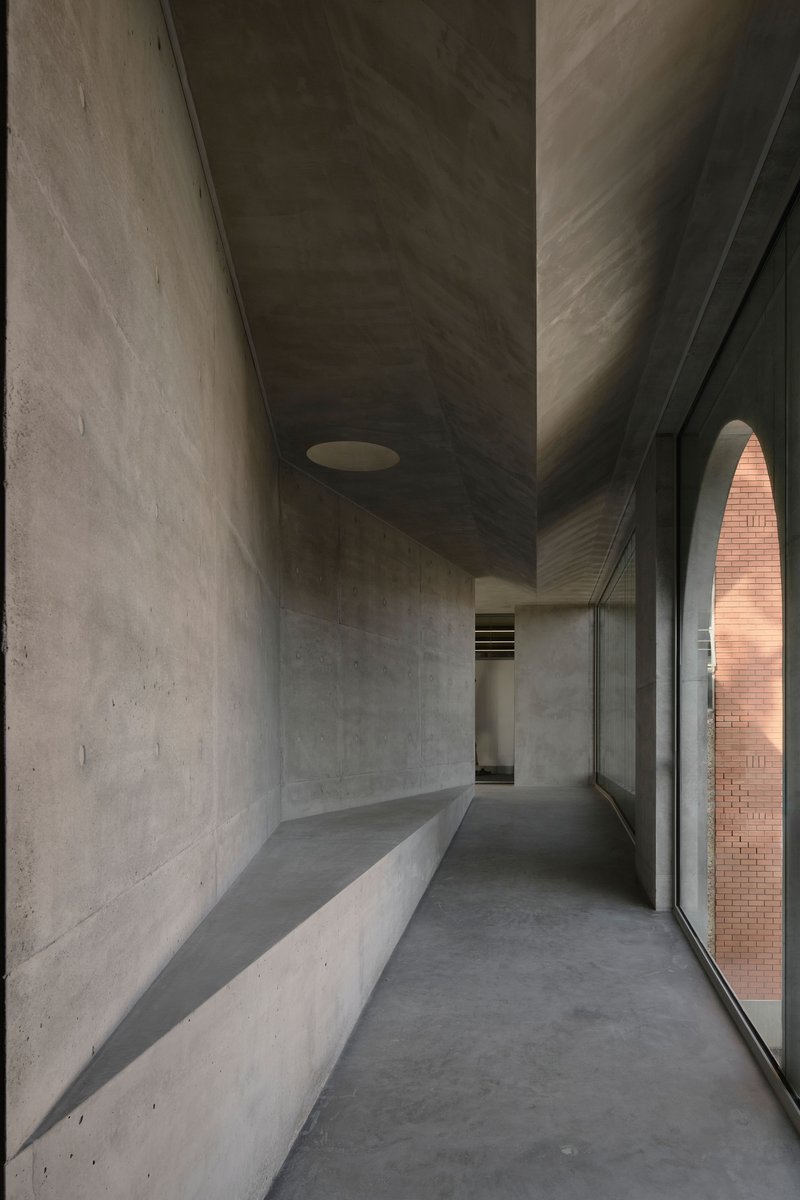Concrete corridor with built-in bench along one wall and full-height glazing overlooking brick courtyard